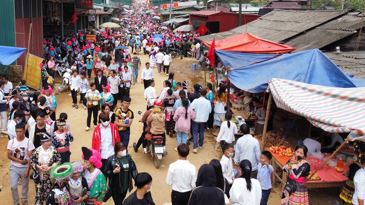 A crowded highland market street with traders in traditional dress and stalls under tarpaulins